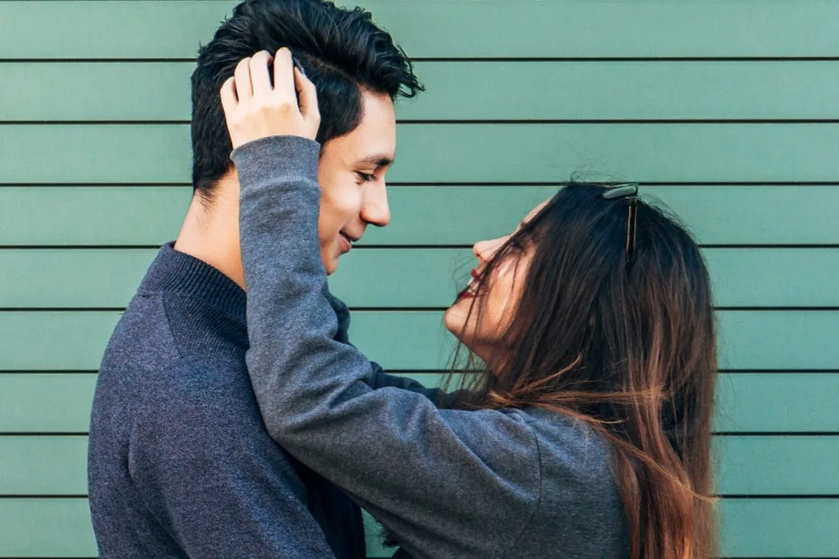 A young couple standing close against a green slatted wall, smiling at each other