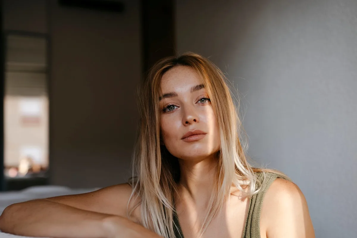 A woman leaning on the edge of a bed in a dim bedroom, looking toward the camera