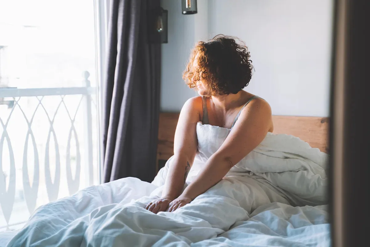 A person sitting alone on an unmade bed, looking out of the window